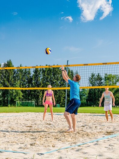 A group of young people is playing beach volleyball on a sand court. The sky is blue with some clouds and the surroundings are green. | © Mias Photoart