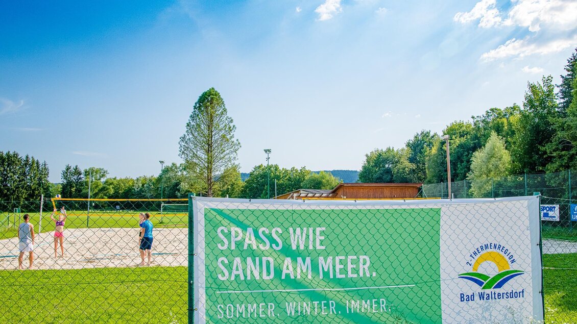 Ein schöner Sandplatz mit Menschen, die Volleyball spielen. Im Hintergrund sind Bäume und ein blauer Himmel zu sehen. | © Mias Photoart