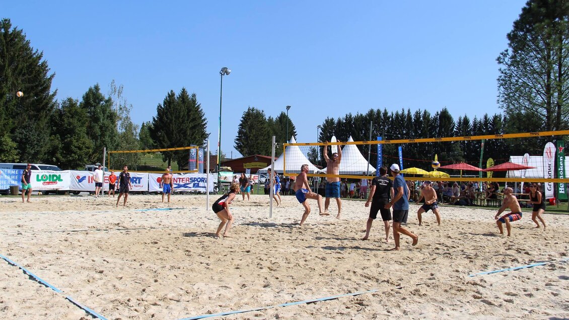 Ein spannendes Beachvolleyballspiel mit mehreren Spielern im Sand. Im Hintergrund sind Stände und Sonnenschirme sichtbar. | © lederer
