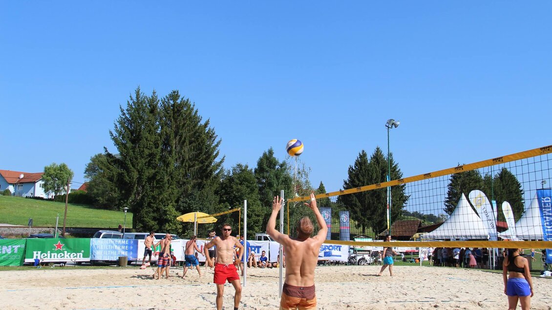 Ein Strandvolleyballspiel unter klarem blauen Himmel. Spieler sind aktiv am Ball, umgeben von Bäumen und Zuschauern. | © lederer