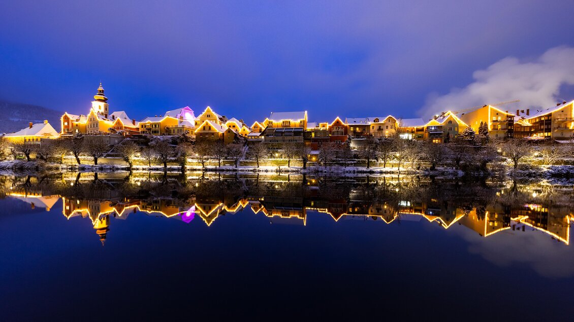 Eine wunderschöne Stadtansicht bei Nacht mit erleuchteten Gebäuden entlang des Wassers. Die Lichter spiegeln sich im ruhigen Wasser und schaffen eine malerische Atmosphäre. | © (c) Region Graz - Harry Schiffer