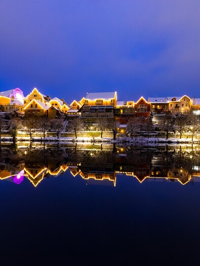 Eine wunderschöne Stadtansicht bei Nacht mit erleuchteten Gebäuden entlang des Wassers. Die Lichter spiegeln sich im ruhigen Wasser und schaffen eine malerische Atmosphäre. | © (c) Region Graz - Harry Schiffer