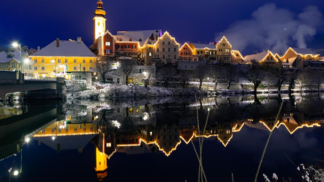 Eine malerische Stadtansicht bei Nacht mit einem beleuchteten Gebäude und einer Uhrturm. Der klare Himmel spiegelt sich im ruhigen Wasser des Flusses. | © (c) Region Graz - Harry Schiffer