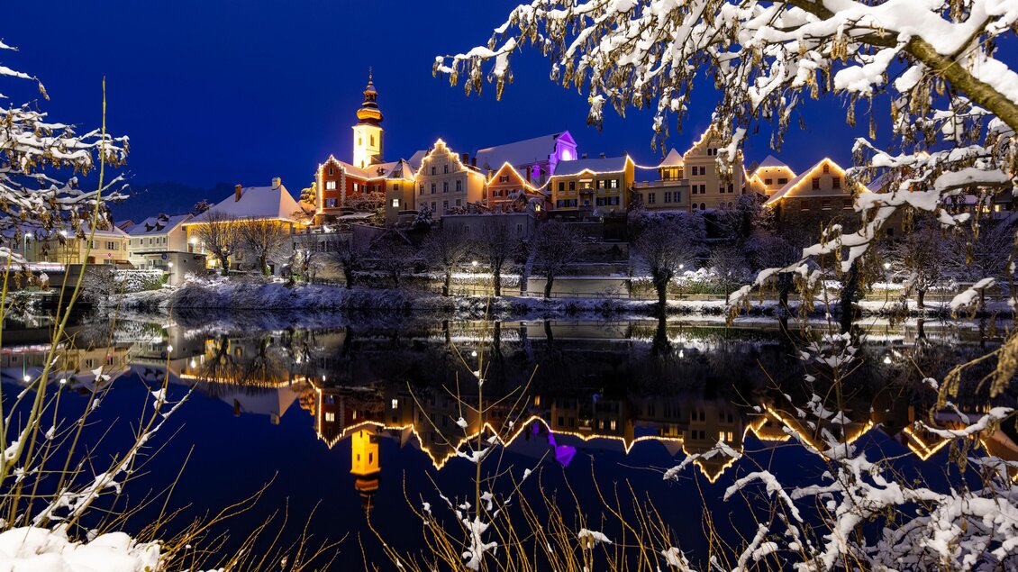 Eine malerische Winterlandschaft mit schneebedeckten Bäumen und einem ruhigen Wasser, das die Lichter eines beleuchteten Gebäudes widerspiegelt. Der Himmel ist dunkelblau und verleiht der Szene eine charmante Atmosphäre. | © (c) Region Graz - Harry Schiffer