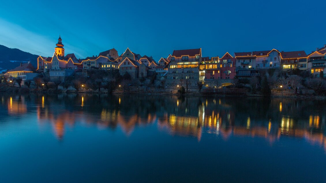 Eine malerische Stadtansicht am Wasser bei Dämmerung, mit beleuchteten Gebäuden und einer ruhigen Spiegelung im Wasser. Der Himmel ist blau mit einem Hauch von Licht. | © (c) Region Graz - Harry Schiffer