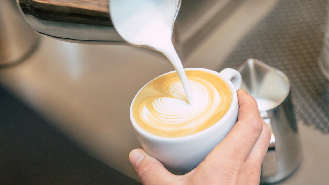 Eine Hand gießt geschäumte Milch in eine Tasse mit Kaffee. Der Kaffee hat eine schöne Latte-Art-Oberfläche. | © Bellaggio