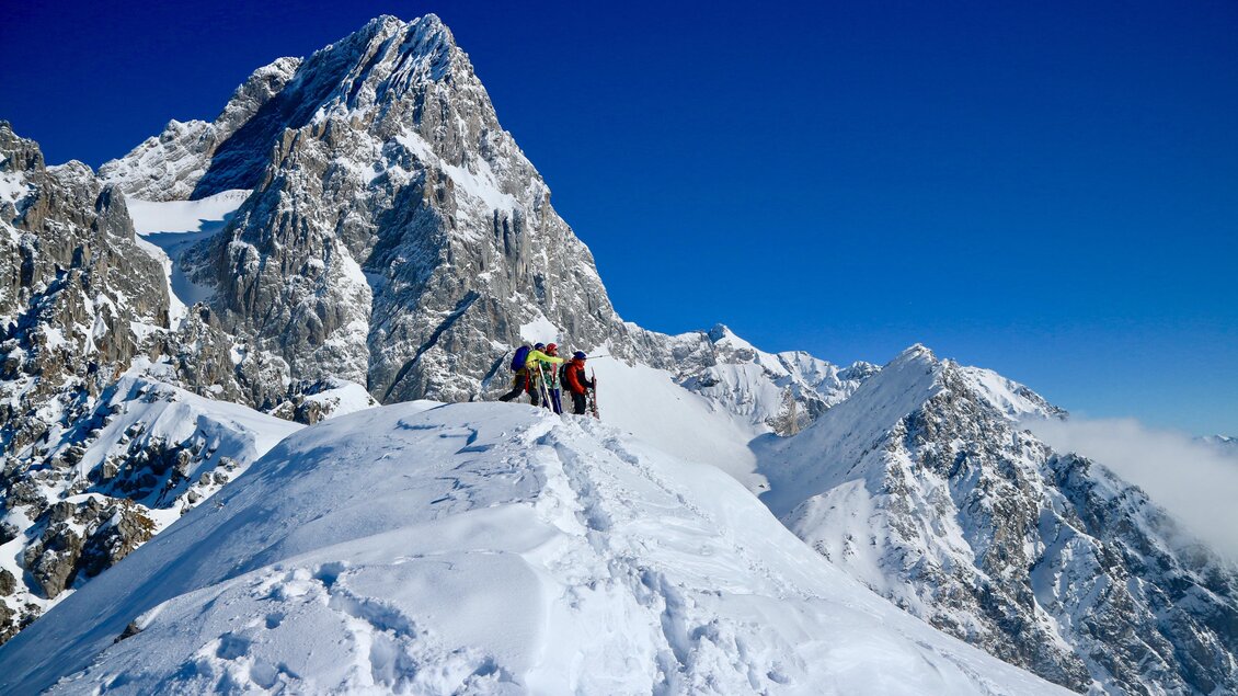 Eine Gruppe von Bergsteigern steht auf einem schneebedeckten Gipfel. Im Hintergrund sind majestätische Berge unter einem klaren blauen Himmel zu sehen. | © Heli Rettensteiner