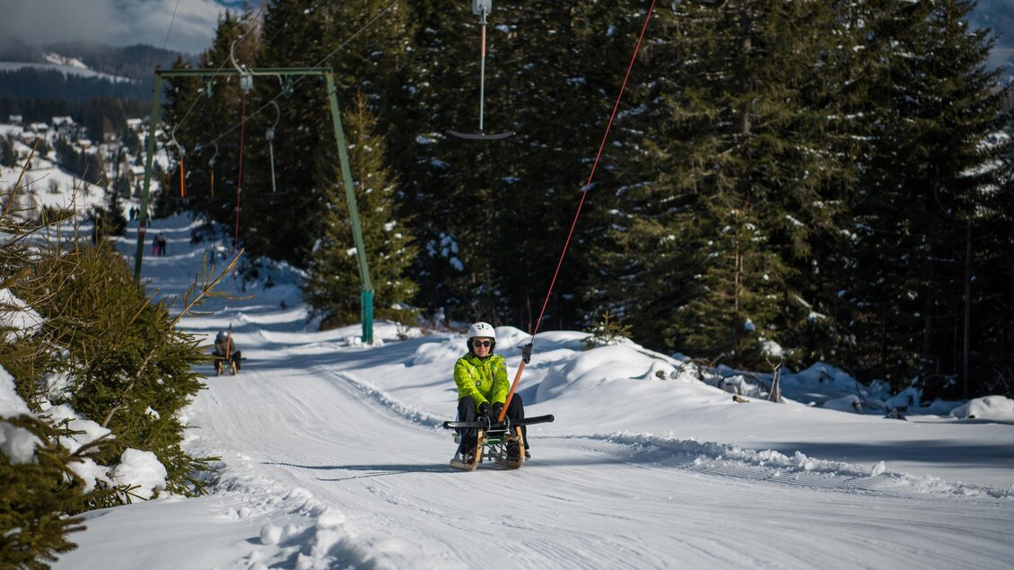 Ein Wintersportler fährt mit einem Schlitten die verschneite Piste hinunter. Im Hintergrund sind schneebedeckte Bäume und die Seilbahn sichtbar. | © Rieseralm