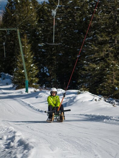 A winter sports enthusiast is sliding down the snowy slope on a sled. In the background, snow-covered trees and the ski lift are visible. | © Rieseralm