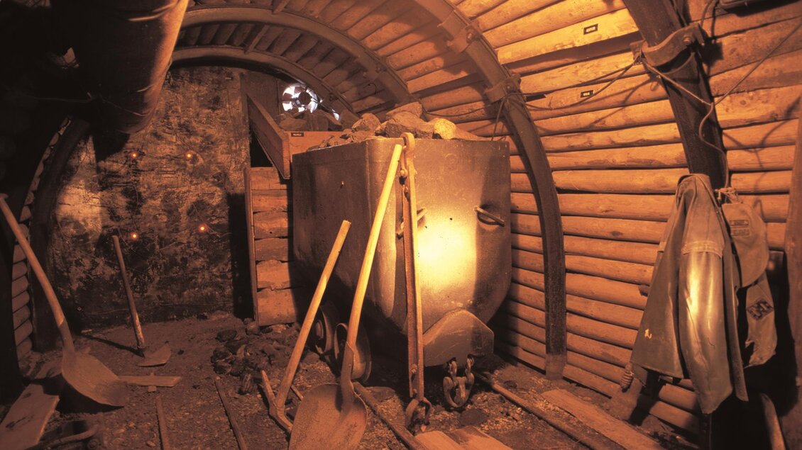 Ein unterirdischer Stollen mit Holzverkleidung und einem Förderwagen. Werkzeuge wie Schaufeln liegen im Raum verstreut. | © Bergbauschaustollen Pölfing Brunn