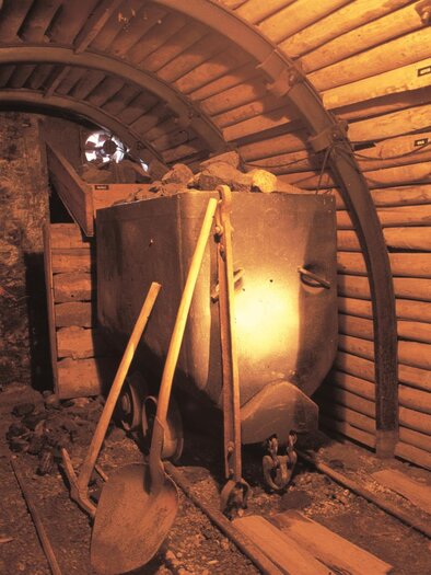 Ein unterirdischer Stollen mit Holzverkleidung und einem Förderwagen. Werkzeuge wie Schaufeln liegen im Raum verstreut. | © Bergbauschaustollen Pölfing Brunn