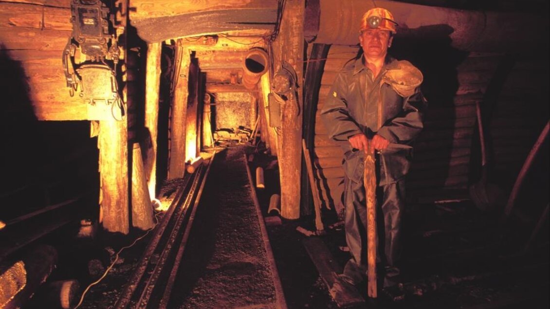 Ein Bergmann steht in einem schmalen Minenschacht. Die Wände sind aus Holz, und schwaches Licht beleuchtet den Tunnel. | © Bergbauschaustollen Pölfing Brunn