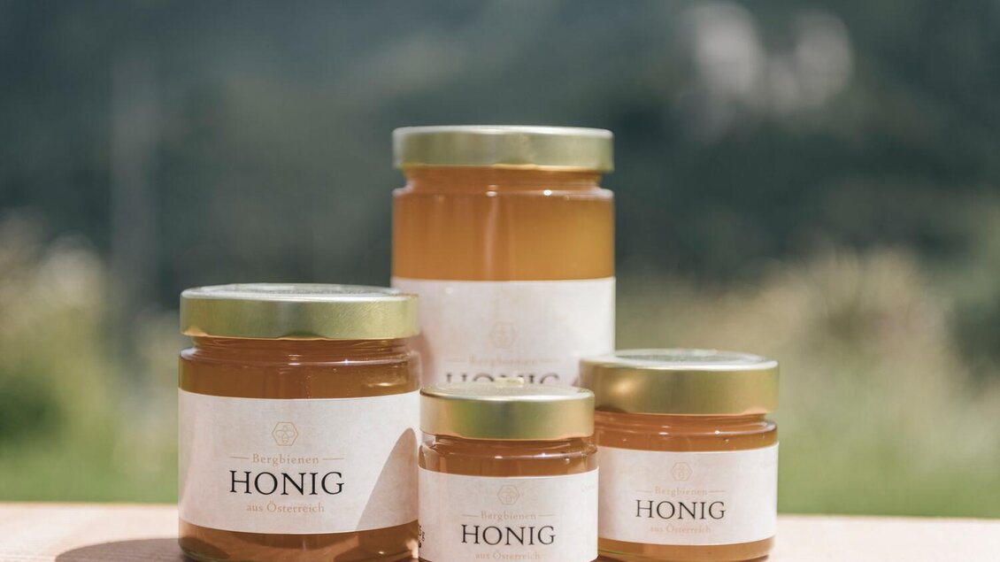 Verschiedene Gläser mit Honig stehen auf einem Holztisch. Im Hintergrund sind sanfte Hügel mit grünem Gras zu sehen. | © Stefan Leitner