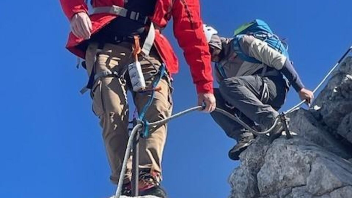 Zwei Bergsteiger klettern an einem Felsen bei klarem blauen Himmel. Der erste Trägt einen roten Helm und eine rote Jacke, während der zweite sich an einem Seil festhält. | © Walter Walcher