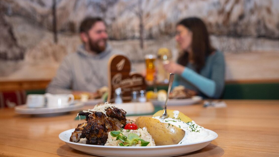 Ein Teller mit leckerem Essen, darunter Fleisch, Kartoffeln und Gemüse, steht im Vordergrund. Im Hintergrund sitzen zwei Personen und genießen eine Mahlzeit in einem gemütlichen Restaurant. | © Harald Steiner