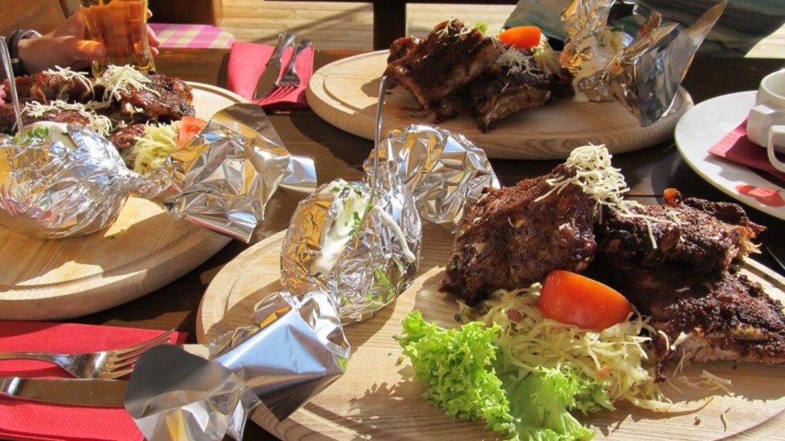 Ein rustikales Gericht mit gegrilltem Fleisch, frischen Gemüsesalaten und Folienkartoffeln auf Holzplatten serviert. Die Sonne scheint auf das köstliche Essen. | © Berggasthof Bottinghaus
