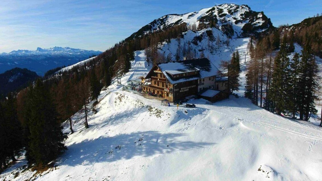 Eine malerische Schnee-Landschaft mit einem rustikalen Gebäude und hohen Bäumen. Im Hintergrund sind schneebedeckte Berge und ein klarer Himmel zu sehen. | © Hollhaus