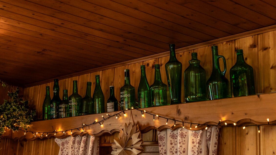 Eine Reihe von grünen Glasflaschen steht auf einem Holzregal. Die Wand ist aus Holz mit einer warmen, rustikalen Atmosphäre. | © Berggasthof Hollhaus, needful.at