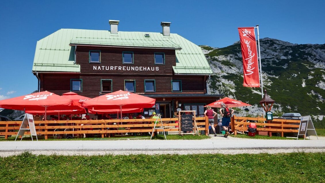 Ein gemütliches Naturfreundehaus mit roten Sonnenschirmen und einer schönen Berglandschaft im Hintergrund. Die Gäste genießen die frische Luft und die Aussicht. | © Karin Klammer Fotografie