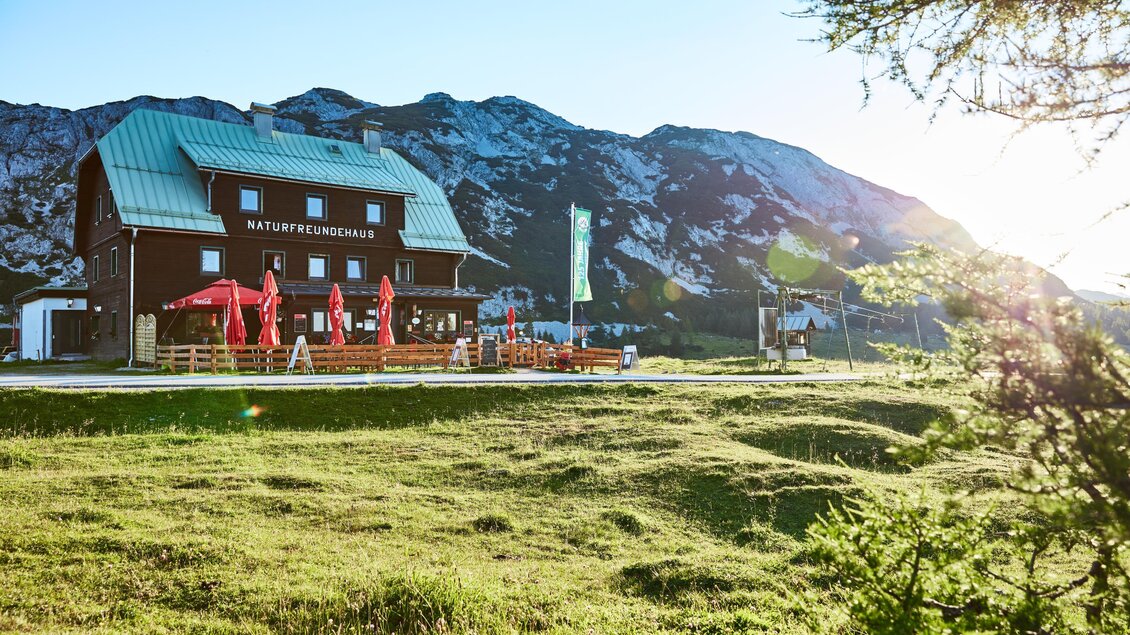 Eine gemütliche Berghütte mit roter Terrasse und Sonnenschirmen. Im Hintergrund sind majestätische Berge zu sehen, und die Sonne strahlt warm über die Landschaft. | © Karin Klammer Fotografie