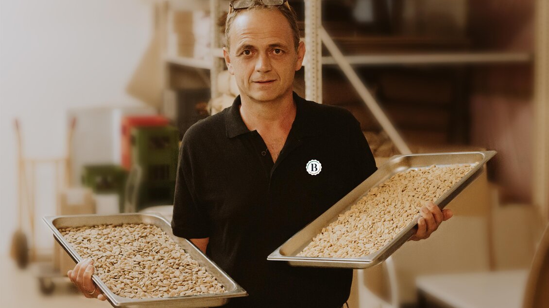 Ein Mann hält zwei Tabletts mit Nüssen in der Hand. Der Hintergrund zeigt eine Werkstatt mit verschiedenen Materialien. | © Berghofer Farmery