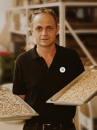 Ein Mann hält zwei Tabletts mit Nüssen in der Hand. Der Hintergrund zeigt eine Werkstatt mit verschiedenen Materialien. | © Berghofer Farmery
