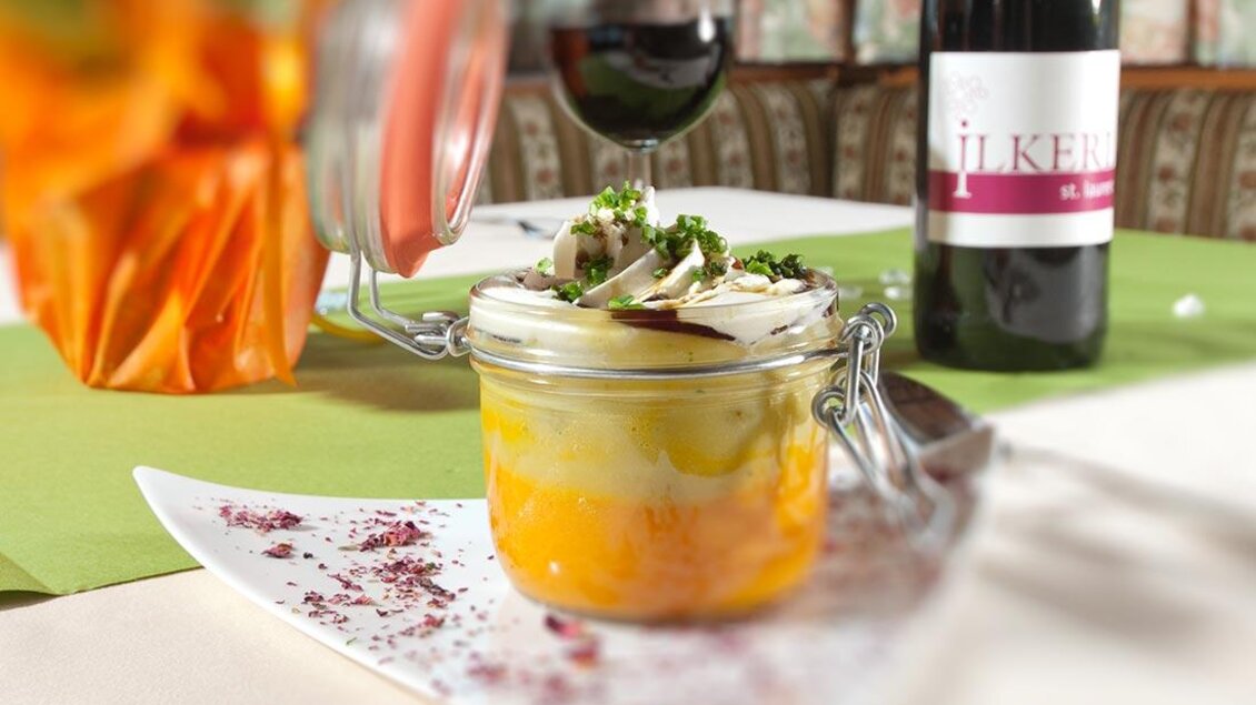 Ein Glas mit einer bunten Vorspeise, das auf einem elegantem Teller serviert ist. Im Hintergrund steht eine Flasche Wein und ein Blumenarrangement. | © Berghotel Türlwand
