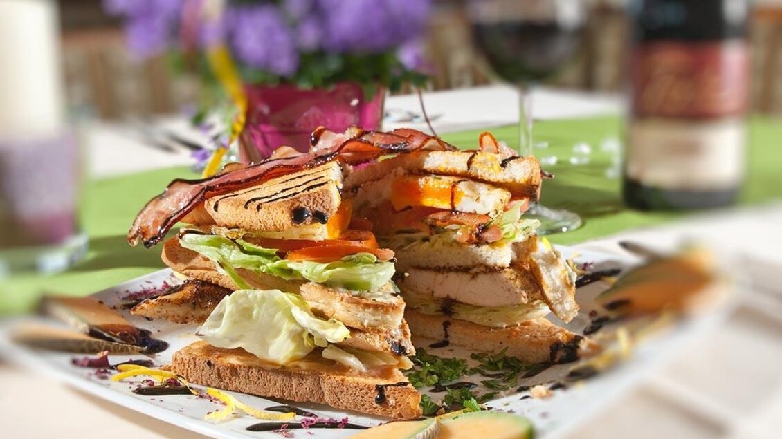 Ein üppiger Sandwich-Turm auf einem Teller, gefüllt mit Schinken, Salat und Hühnerfleisch. Im Hintergrund sind Wein und eine blühende Pflanze zu sehen. | © Berghotel Türlwand