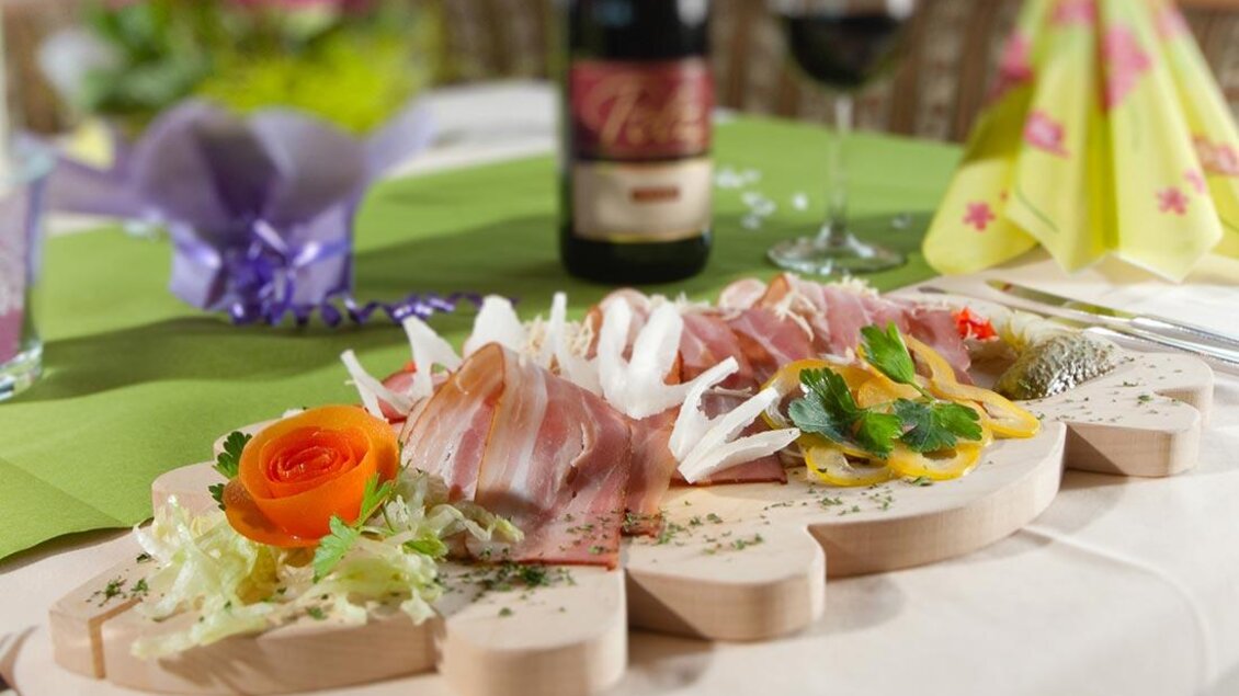 Ein appetitliches Platt mit Aufschnitt und frischem Gemüse auf einem Holzbrett. Im Hintergrund sind ein Glas Rotwein und dekorierte Tischsets zu sehen. | © Berghotel Türlwand