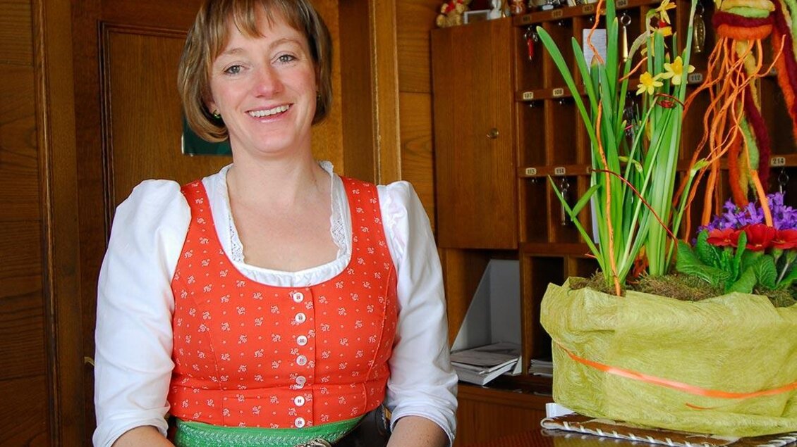Eine freundliche Frau in traditioneller Tracht lächelt am Empfang. Im Hintergrund sind dekorative Pflanzen und Holzverzierungen zu sehen. | © Berghotel Türlwand