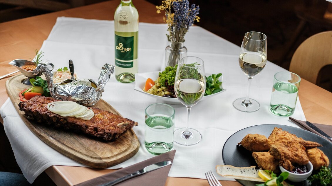 Ein schön gedeckter Tisch mit zwei Tellergerichten, Wein und einer Salatplatte. Im Vordergrund liegt ein Stück Fleisch auf einem Holzbrett, umgeben von Gemüse und Getränken. | © Bergstadl