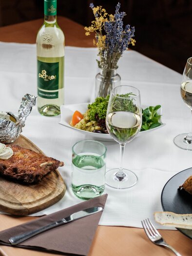 Ein schön gedeckter Tisch mit zwei Tellergerichten, Wein und einer Salatplatte. Im Vordergrund liegt ein Stück Fleisch auf einem Holzbrett, umgeben von Gemüse und Getränken. | © Bergstadl