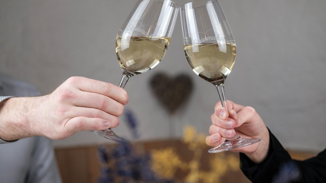 Zwei Gläser mit weißen Wein stoßen an. Im Hintergrund sind dekorative Elemente sichtbar. | © Bergstadl
