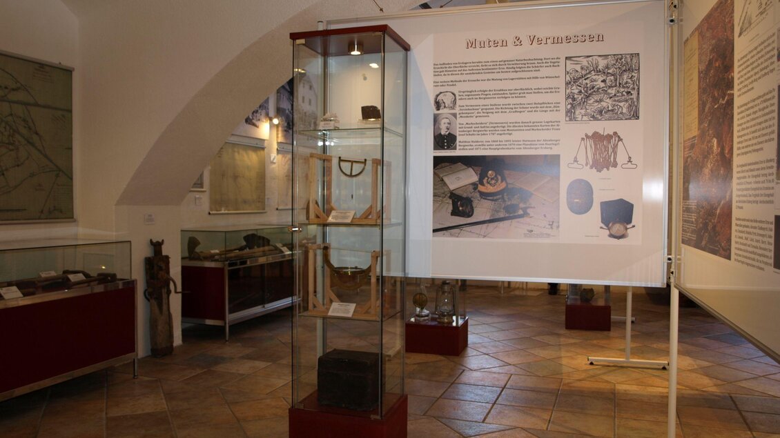 Ein Ausstellungsraum mit Vitrinen und historischen Artefakten. An den Wänden sind informative Tafeln mit Texten und Bildern zu sehen.