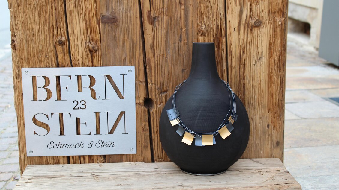 Eine schwarze Vase mit einem auffälligen Halsband steht auf einer Holzplatte. Im Hintergrund ist ein Schild mit der Aufschrift "BERN 23 STEIN" zu sehen. | © Bernstein 23