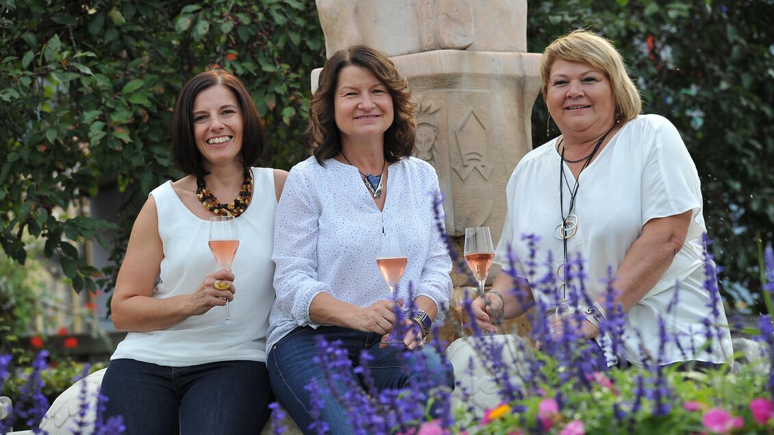 Drei Frauen sitzen lächelnd im Freien und halten Gläser mit Sekt. Im Hintergrund sind bunte Blumen und eine Statue zu sehen. | © Bernstein 23