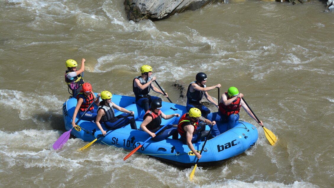 Eine Gruppe von Menschen paddelt in einem blauen Rafting-Boot auf einem reißenden Fluss. Alle tragen Helme und Schwimmwesten. | © BAC