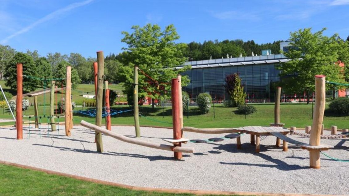 Ein Spielplatz mit Holzelementen und einem Kiesboden. Im Hintergrund sind Bäume und ein modernes Gebäude zu sehen. | © Robert Cescutti