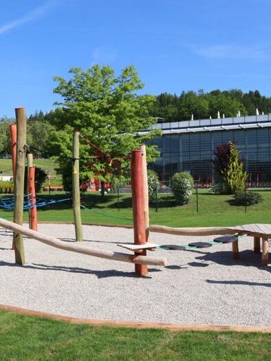 Ein Spielplatz mit Holzelementen und einem Kiesboden. Im Hintergrund sind Bäume und ein modernes Gebäude zu sehen. | © Robert Cescutti