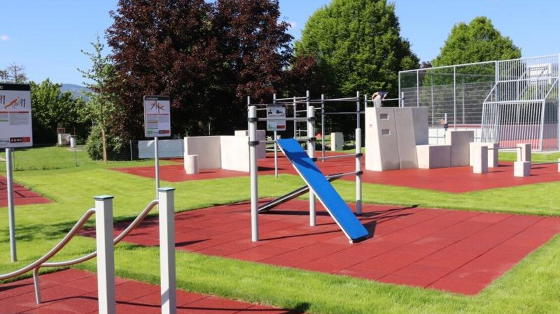 Ein moderner Fitnessbereich im Freien mit verschiedenen Geräten und einem Spielgerät. Der rote Gummiboden sorgt für Sicherheit und Komfort. | © Robert Cescutti