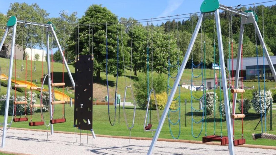 Ein Spielplatz mit Schaukeln und einem Klettergerüst. Im Hintergrund sind grüne Bäume und eine Rutsche zu sehen. | © Robert Cescutti