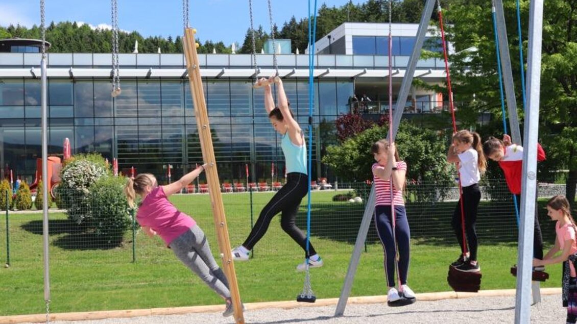 Ein belebter Spielplatz mit Kindern, die an Schaukeln spielen. Im Hintergrund sind grüne Bäume und ein modernes Gebäude zu sehen. | © Robert Cescutti