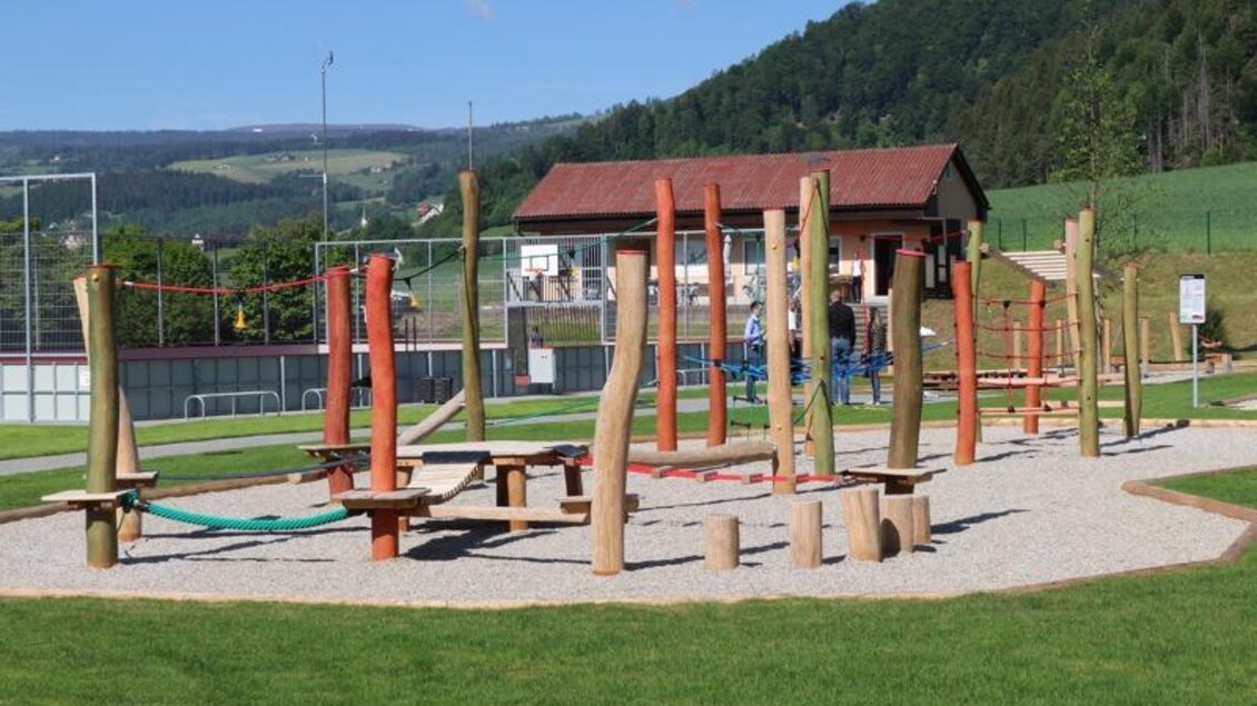 Ein moderner Spielplatz mit Kletterelementen aus Holz. Im Hintergrund ist ein Gebäude und grüne Wiesen zu sehen. | © Robert Cescutti