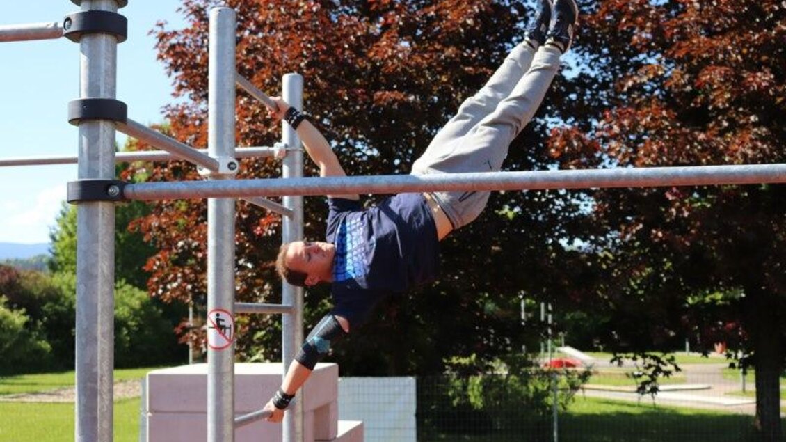 Ein junger Mann macht eine akrobatische Übung an einem Trainingsgerät im Freien. Im Hintergrund sind Bäume und ein klarer Himmel zu sehen. | © Robert Cescutti