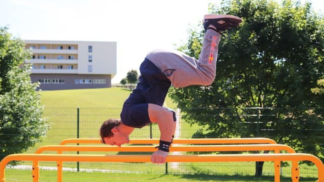 Ein Mann тренiert an einer orangefarbenen Trainingsstation. Er ist in einer Hüftstandposition mit gestreckten Beinen und einem konzentrierten Gesichtsausdruck. | © Robert Cescutti