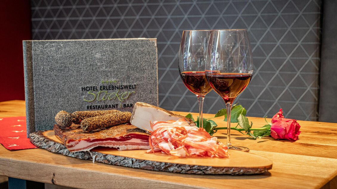 Ein Holzbrett mit einer Auswahl an Wurst- und Käsesorten, begleitet von zwei Gläsern Rotwein. Im Hintergrund steht ein elegantes Menübuch des Hotels. | © c.hoeflehner