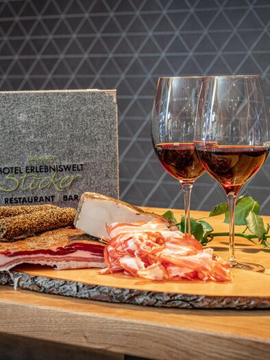 A wooden board with a selection of sausages and cheeses, accompanied by two glasses of red wine. In the background, there is an elegant menu book of the hotel. | © c.hoeflehner