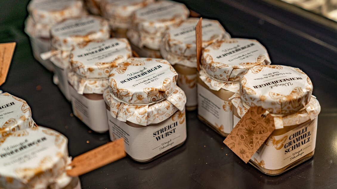 Eine Reihe von Gläschen mit verschiedenen Aufstrichen steht auf einer Präsentationsfläche. Die Etiketten zeigen kreative Namen wie "Streichwurst" und "Chili-Gemüse-Schmalz". | © weingut schneeberger