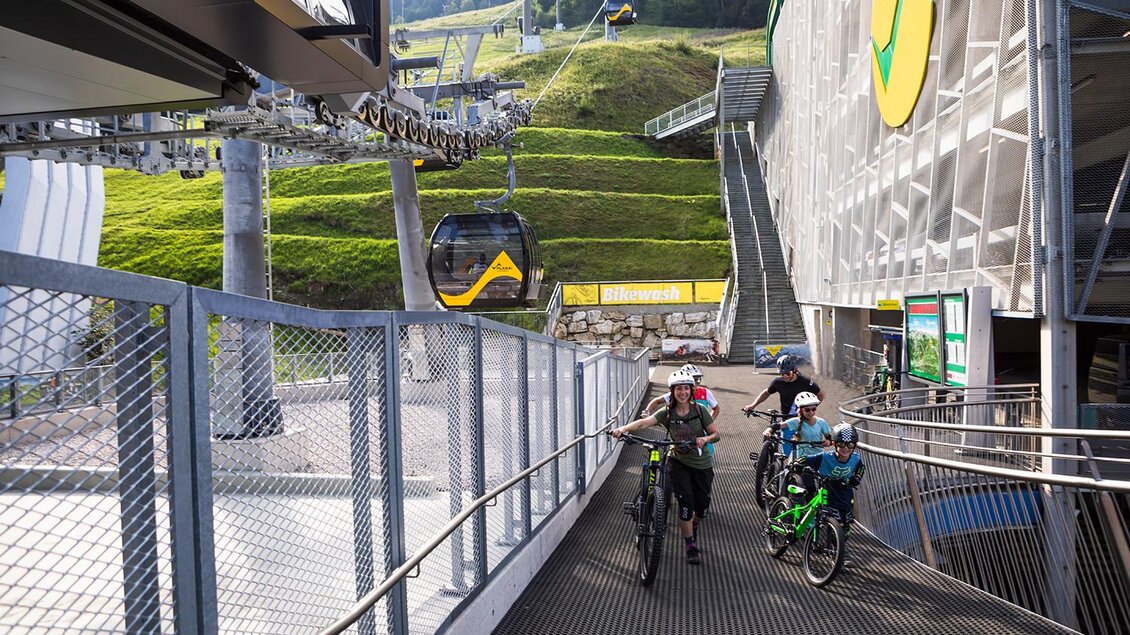 Eine Seilbahnstation in den Bergen mit Radfahren auf der Rampe. Im Hintergrund sind grüne Wiesen und Wanderrouten zu sehen. | © Roland Haschka