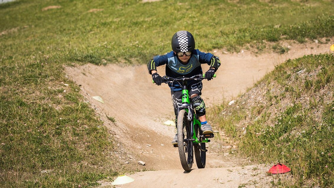 Ein Junge fährt mit einem Mountainbike über einen Dirtjump. Die Umgebung ist grün und sonnig, ideal für Outdoor-Aktivitäten. | © Roland Haschka